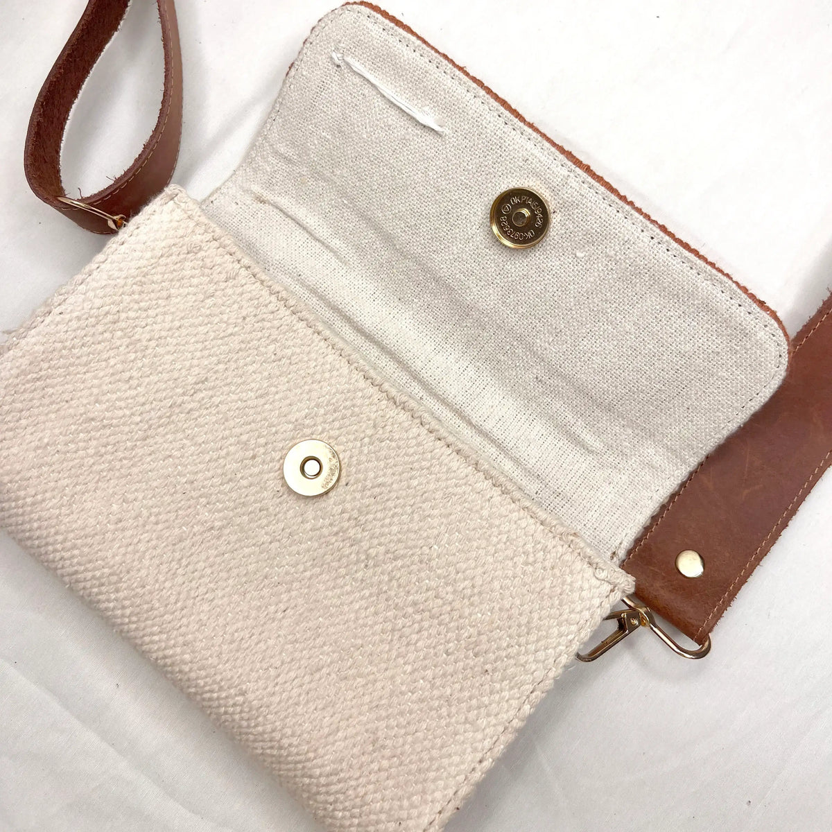 An open handbag is presented on a light background, revealing its interior lined with cream fabric. The bag features a magnetic snap closure and a brown leather strap, showcasing its minimalist design and functionality.