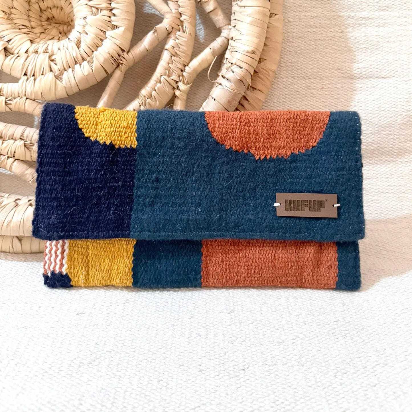 This image shows a clutch. The clutch has a geometric pattern in shades of navy blue, mustard yellow, and burnt orange. The front of the clutch has a small leather logo tag on it. The clutch appears to be made of a plush, textured material, likely yarn or thread. The image is against a plain white background, highlighting the vibrant colors and pattern of the unique handmade accessory.