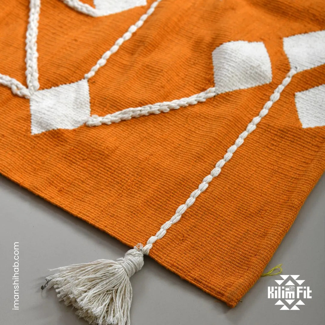 This 1 x 1.5 meter rug features a bold, orange background and a striking white geometric pattern that consists of interconnected diamond shapes, creating a visually engaging composition. The edges are adorned with fringed tassels made of the same material, contributing to the traditional and artisanal aesthetic.