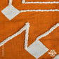 This 1 x 1.5 meter rug features a bold, orange background and a striking white geometric pattern that consists of interconnected diamond shapes, creating a visually engaging composition. The edges are adorned with fringed tassels made of the same material, contributing to the traditional and artisanal aesthetic.