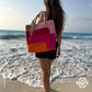 "A person standing on the beach, holding a colorful tote bag with pink, orange, and red stripes, with waves gently lapping at their feet."