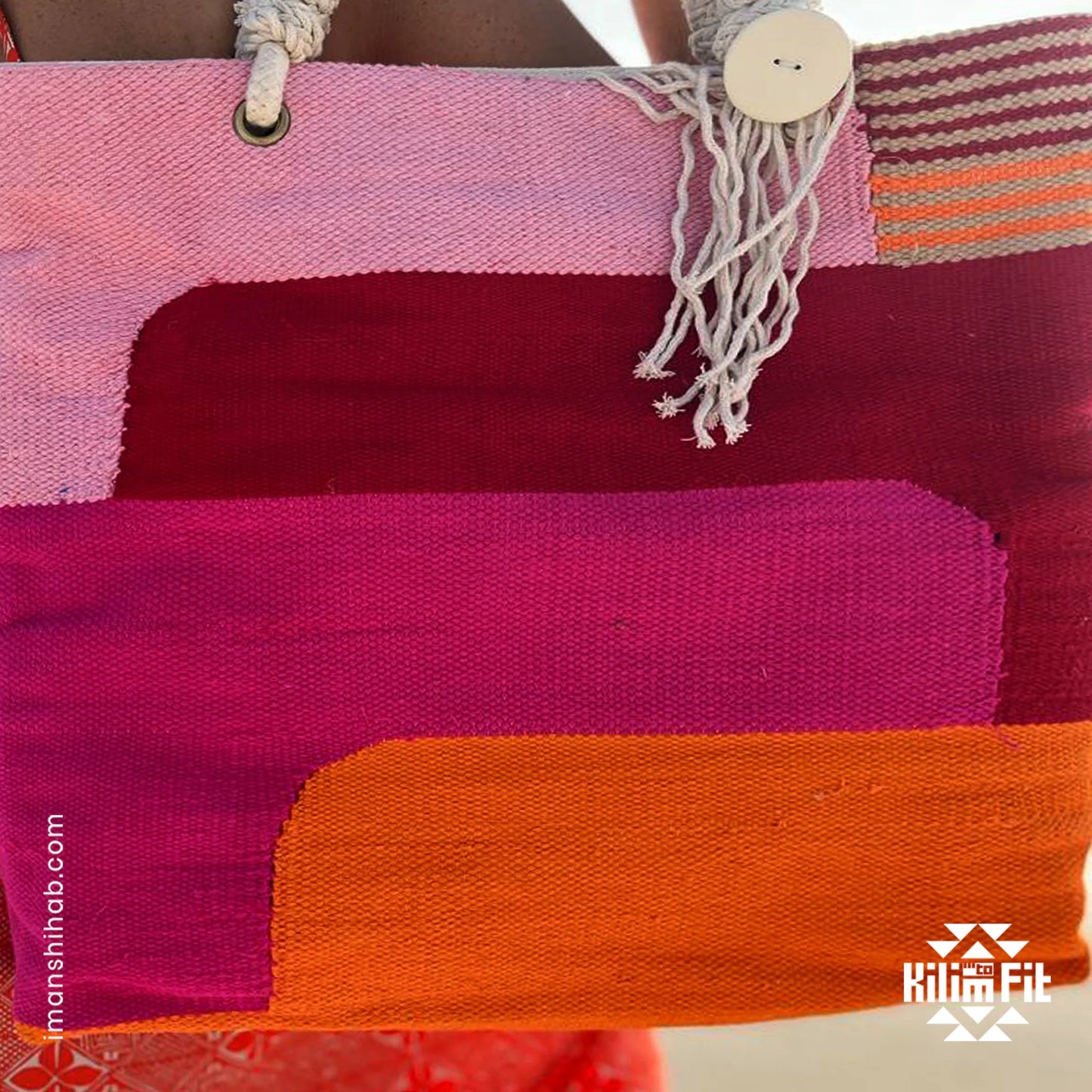 "A close-up of a colorful woven tote bag featuring horizontal stripes in shades of pink, red, and orange. The bag has rope handles with a large button and tassel detail."