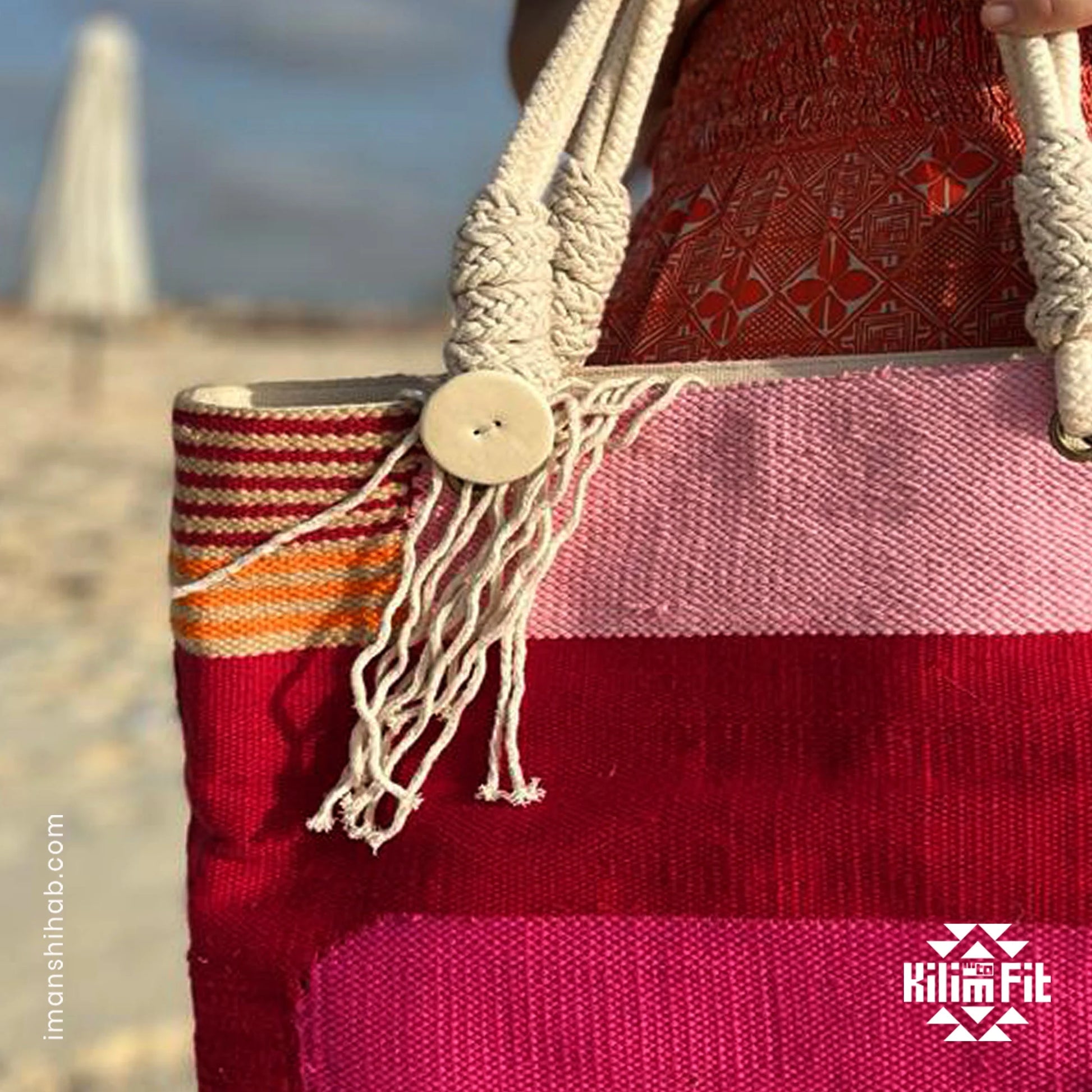 "A close-up of a vibrant, handmade tote bag featuring bold stripes in pink, orange, and red, held by a person on a beach with soft sand in the background. The bag is adorned with a decorative button and macramé detailing."