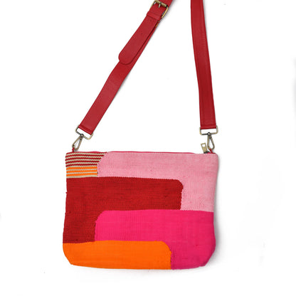 A colorful crossbody bag featuring a patchwork design with shades of pink, orange, red, and yellow. It has a red strap and a textured surface, showcasing various geometric patterns.