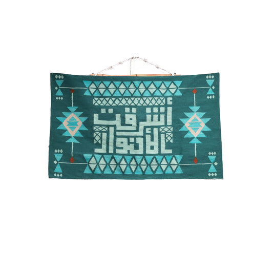 "A decorative wall hanging with Arabic calligraphy in the center, surrounded by geometric patterns in shades of green and mint, displayed on a white wall."