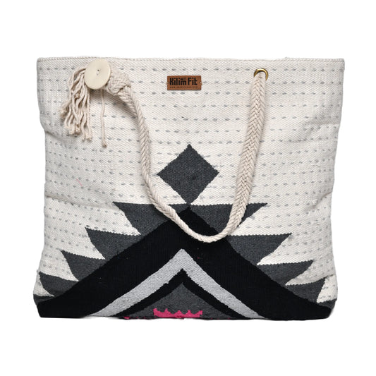 "Front view of a handwoven tote bag with a geometric pattern in black, gray, white, and pink colors. The bag has rope handles and a button closure with a tassel."