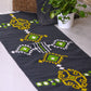 The image shows a dark gray rug with a geometric pattern in shades of green and yellow. The pattern is composed of repeating motifs, including stylized flowers or medallions, diamonds, and other abstract shapes. The rug has a hand-woven or hand-tufted look, with slightly uneven edges and a visible texture. The rug is lying on a light-colored tile floor, and a potted plant and a woven basket are visible in the background. The overall style is modern with ethnic or tribal influences.