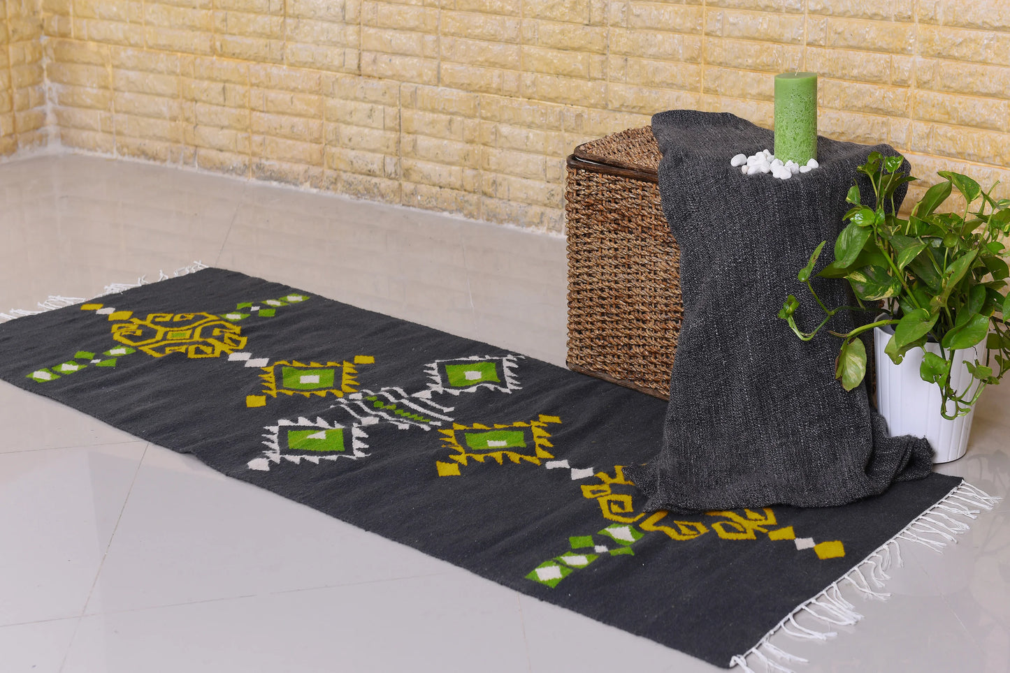 A long, dark rug with geometric patterns in yellow and green is laid on a glossy tiled floor. Nearby, there's a woven storage basket and a small green candle on a table draped with a dark cloth. A potted plant adds a touch of greenery to the scene. The background features a light-colored brick wall.