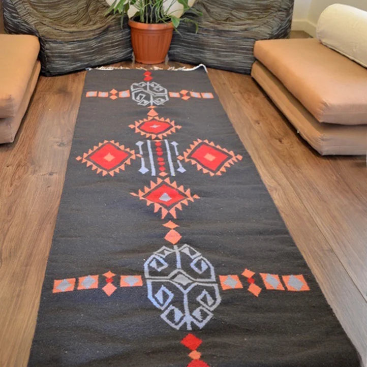 The image shows a long, decorative rug with a black background featuring intricate geometric patterns in red, gray, and white. The rug is placed on a wooden floor and is flanked by two low, beige seating cushions. In the background, there is a potted plant adding a touch of greenery to the setting.