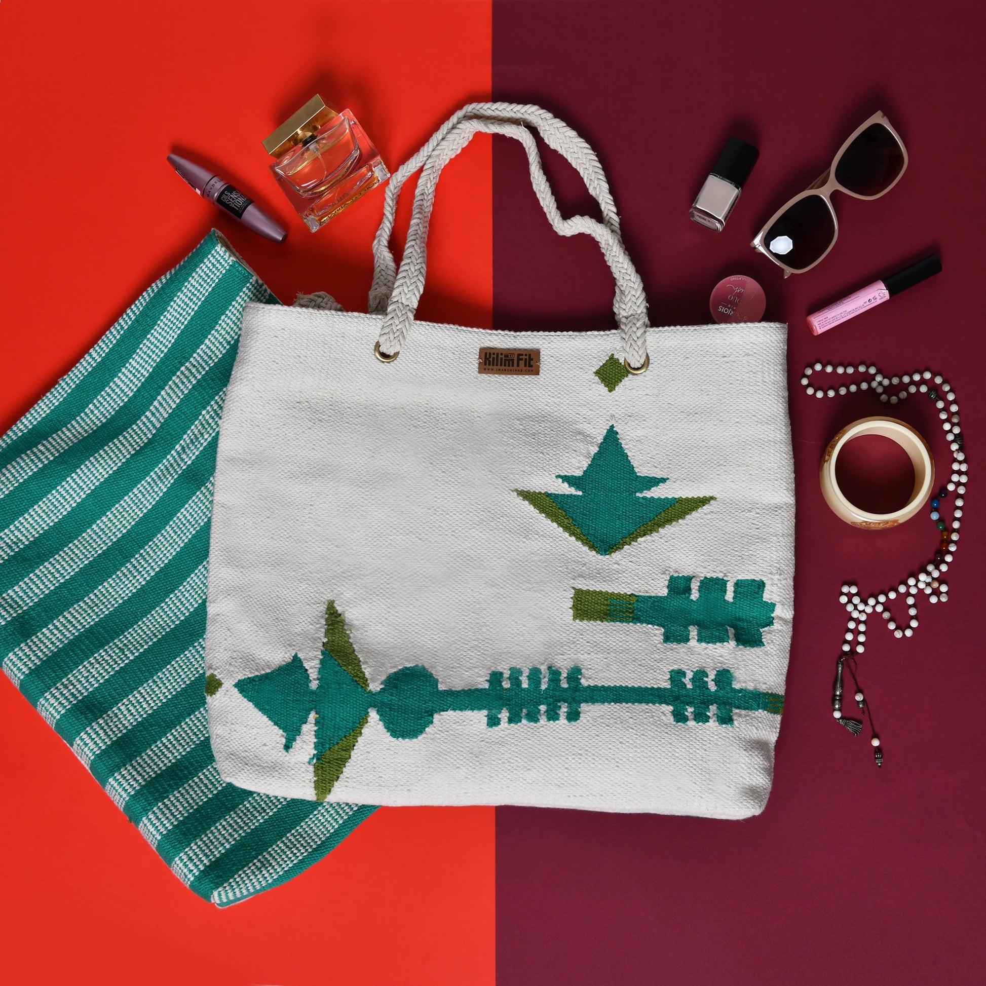 A large, white woven tote bag with green geometric patterns is displayed against a split background of red and deep maroon. Surrounding the bag are various items including a bottle of perfume, several nail polish bottles, a pair of sunglasses, and jewelry such as a bracelet and a necklace. A smaller striped bag in teal and white is also visible.