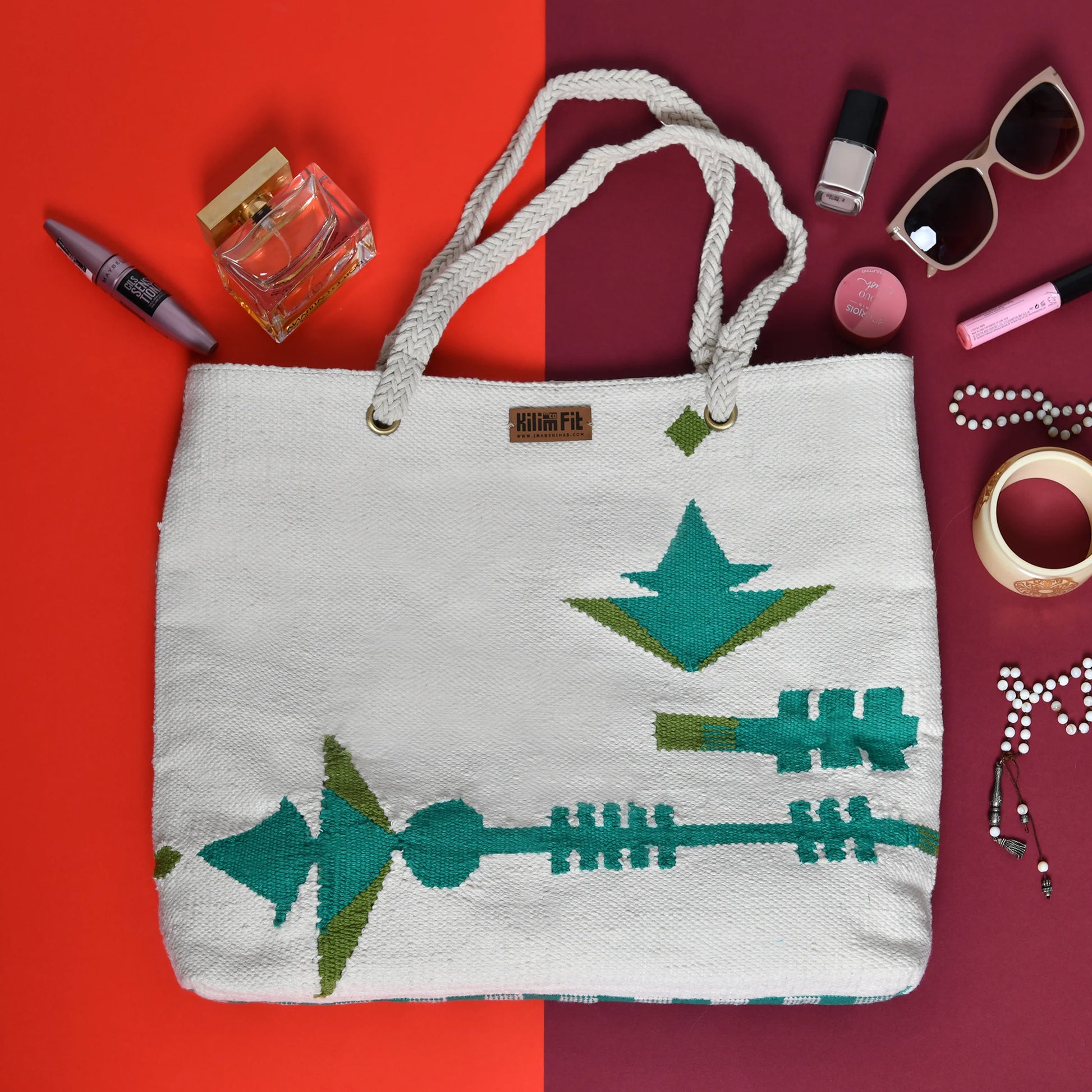 A stylish woven tote bag featuring a geometric design in teal and green hues. The bag is placed on a split background with one side in red and the other in burgundy. Surrounding the bag are various accessories, including a bottle of perfume, nail polish, sunglasses, lipstick, a bracelet, and a necklace, adding a chic touch to the overall composition.