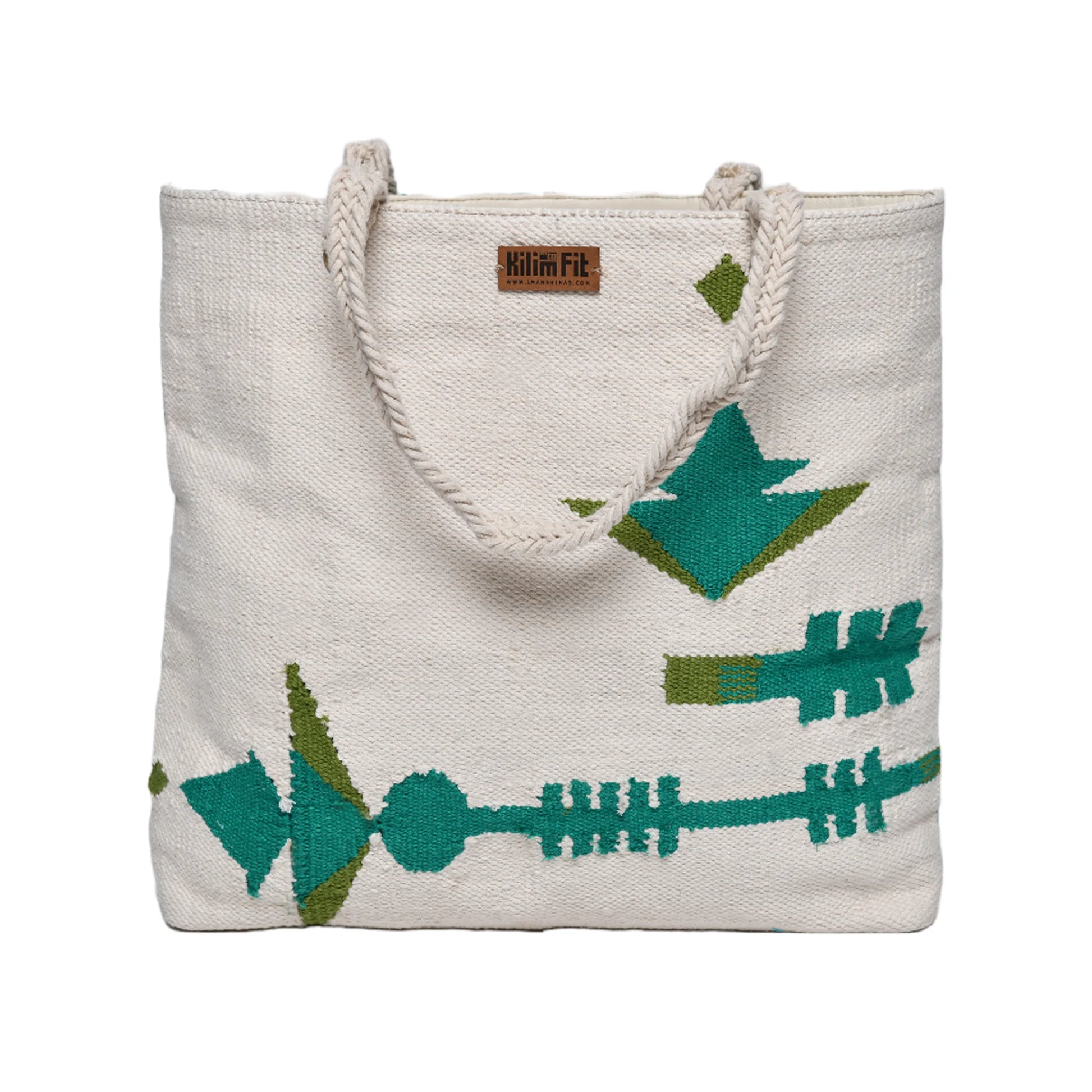 This is an image of a large, woven tote bag featuring a cream-colored background with green and teal geometric patterns. The design includes stylized arrows and figures, giving it a distinctive and artistic look. The bag has sturdy rope handles and a small label at the top that reads "Kilim Fit.