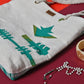 A woven tote bag featuring a design with green arrows and guitar silhouettes. It has a sturdy rope handle and is placed on a colorful background with a decorative ring and a beaded necklace nearby. The overall aesthetic is vibrant and artistic.