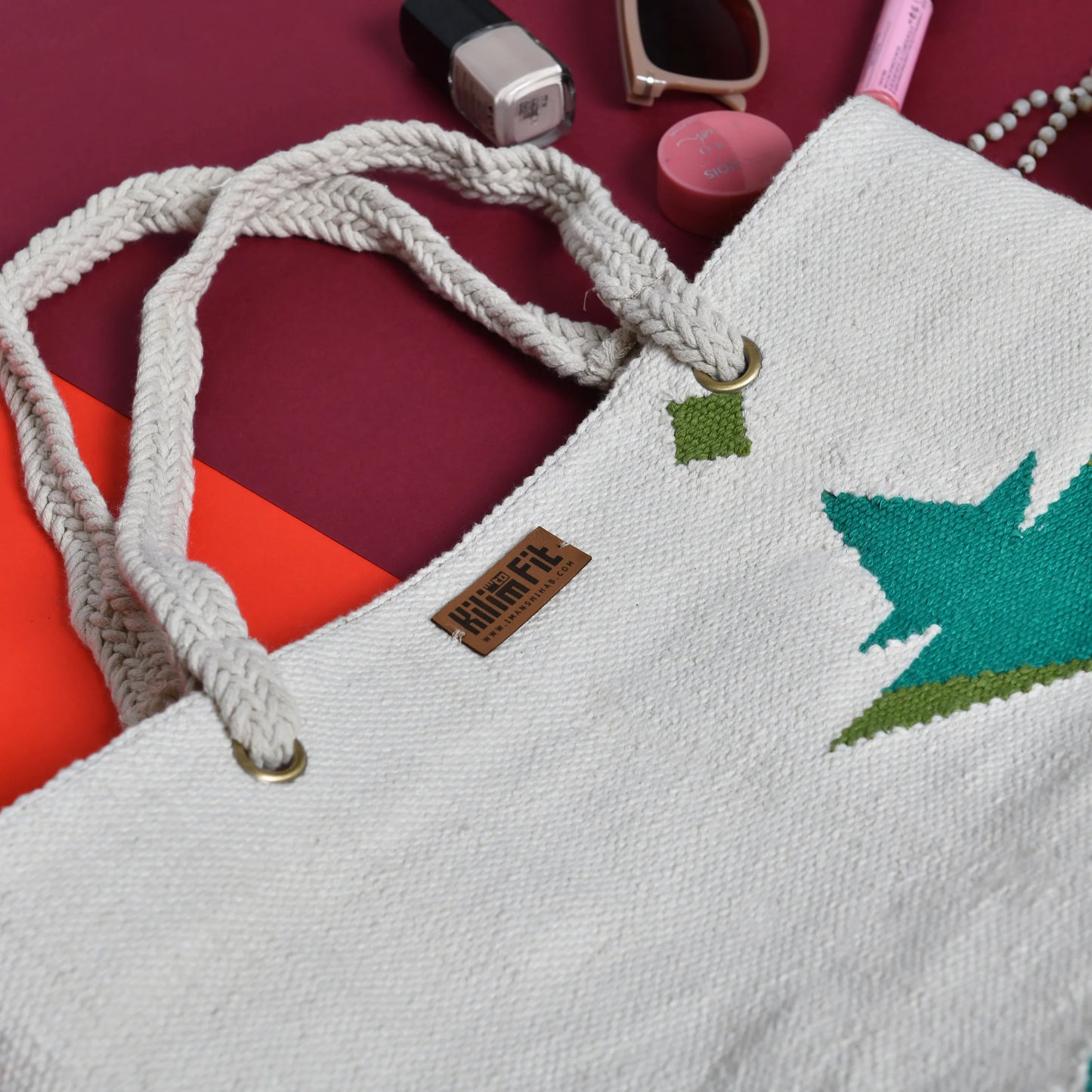 A close-up of a white tote bag with braided handles, featuring a green leaf design and a small brown label that reads "KilimFit." Surrounding the bag are various items, including nail polish, sunglasses, and makeup products, set against a colorful background of red and orange.