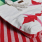 The image features a close-up of a textile item, possibly a tote bag, with a textured surface. The bag is primarily white with vibrant red leaf patterns, and it has a decorative tassel and a button. In the background, there is a striped fabric with red and white lines that contrasts with the bag's design. The overall aesthetic is colorful and handmade.