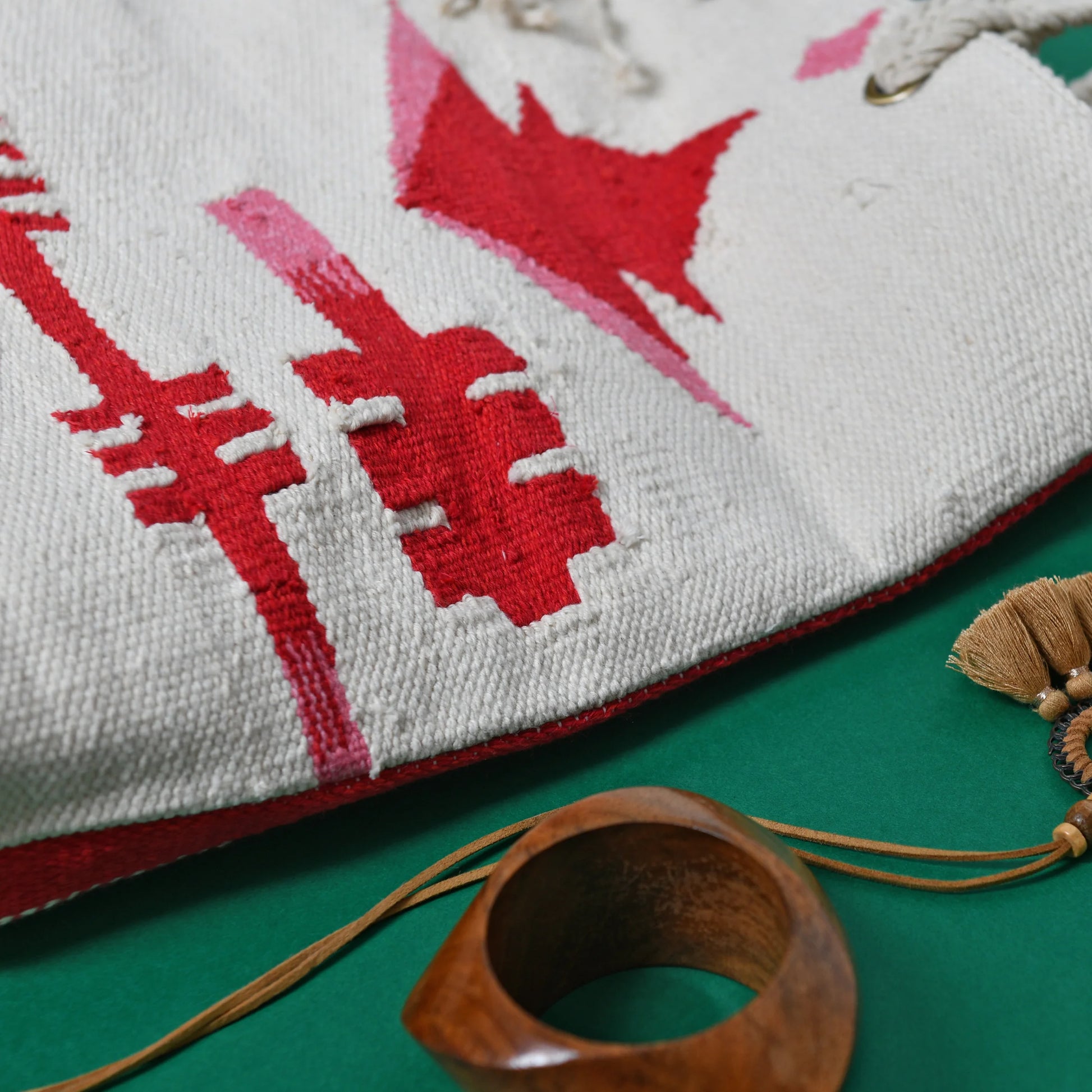 This image features a close-up of a textile or woven bag with a predominantly white background adorned with red abstract patterns, including shapes resembling arrows or lines. The bag is partially open, revealing its inner fabric. Nearby, there are decorative items, including a wooden ring and a tassel with a beige-colored fringe, all set against a green background.