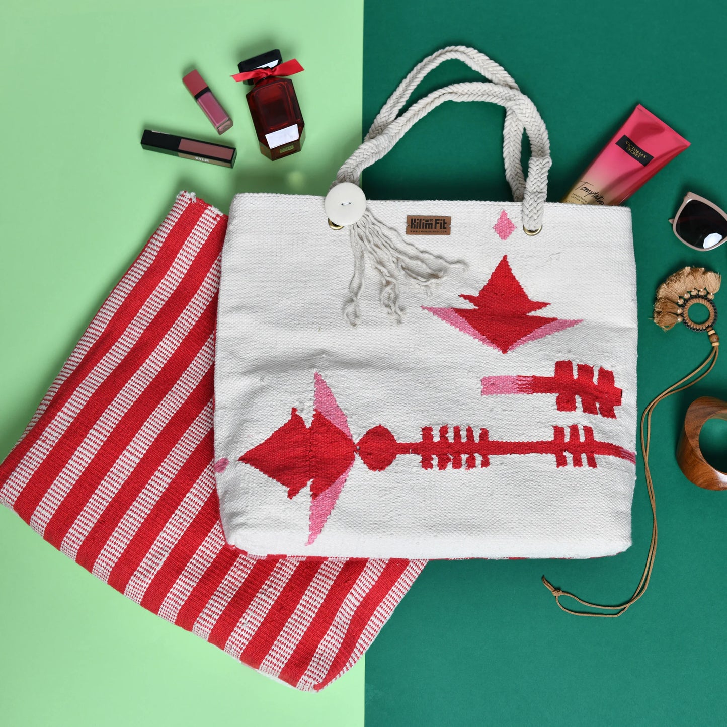 The image features a stylish, woven tote bag with a white background adorned with bold red geometric patterns. It is placed on a split background of green and light green. Next to the bag, there are various beauty products, including lipsticks, nail polish, and a tube of cream, as well as a pair of sunglasses and some accessories like bracelets. A striped fabric in red and white is also visible, complementing the tote bag.