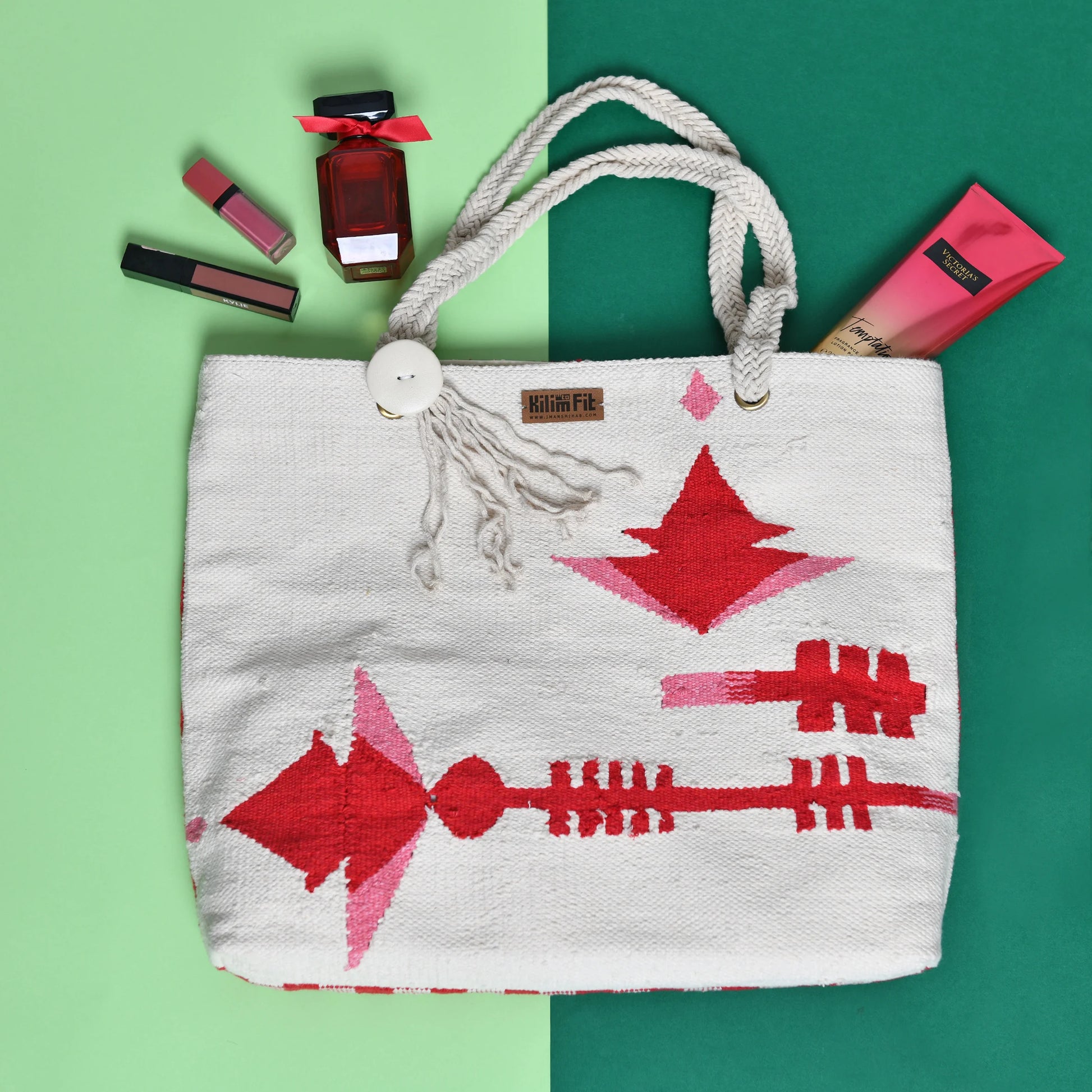 The image shows a stylish, woven tote bag with a colorful design featuring red arrows and geometric shapes. The bag is accompanied by beauty products, including lipsticks and a perfume bottle, arranged around it on a split background of green and a lighter shade. The bag has a button closure and braided handles, adding to its aesthetic appeal.