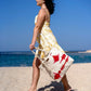 A woman wearing a yellow floral dress walks along a sandy beach, holding a large, white tote bag decorated with red designs. The ocean is visible in the background under a clear blue sky.