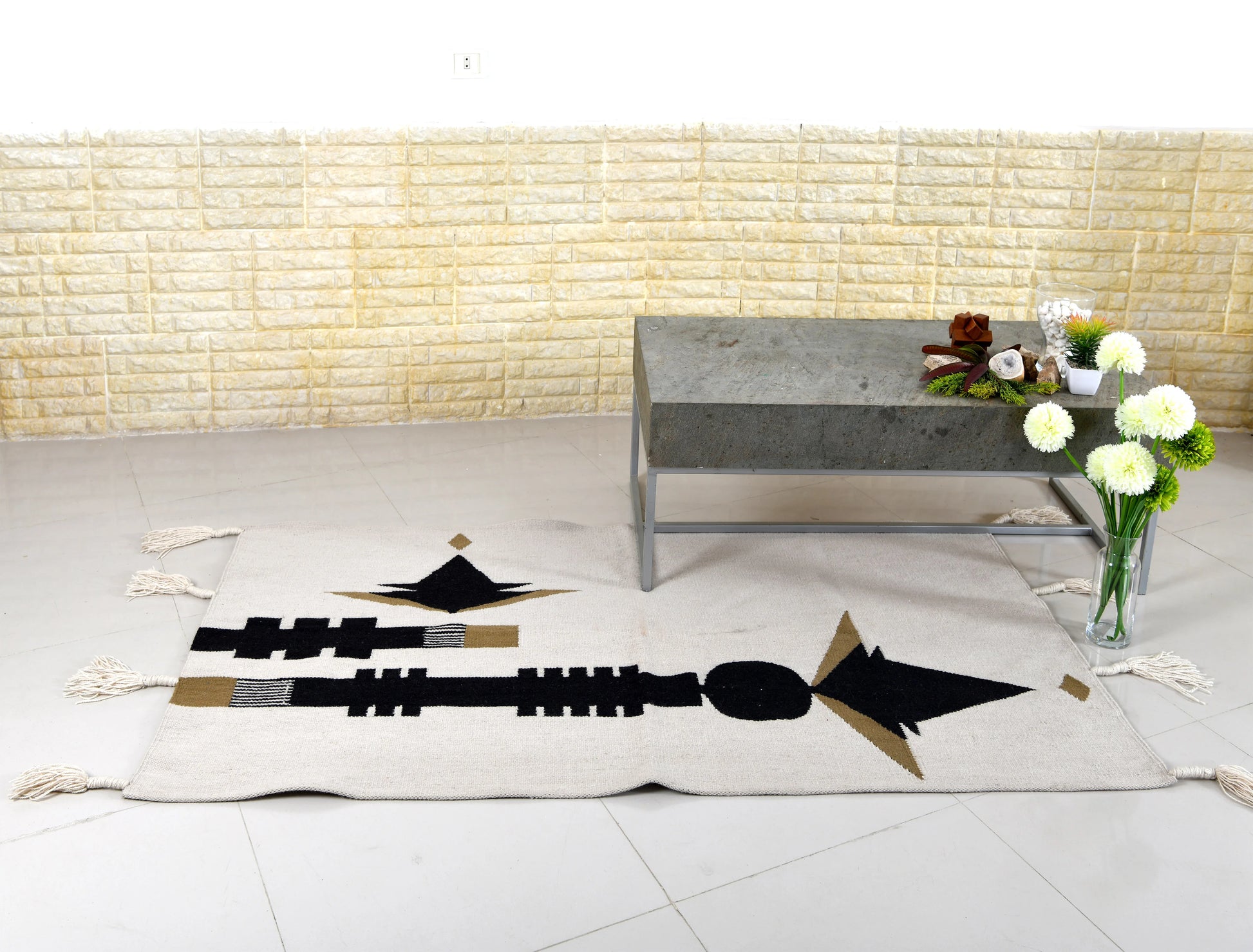 A stylish living room setup featuring a patterned area rug with geometric designs in black and beige. There is a modern, minimalist coffee table made of gray stone positioned on the rug. A vase with white flowers is placed on the table, and the background consists of textured light-colored walls.