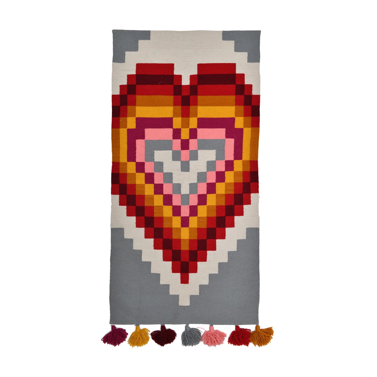 A rectangular rug featuring a bold, colorful heart design made up of various geometric shapes in shades of red, pink, orange, and gray. The bottom of the rug is adorned with colorful tassels in matching hues.