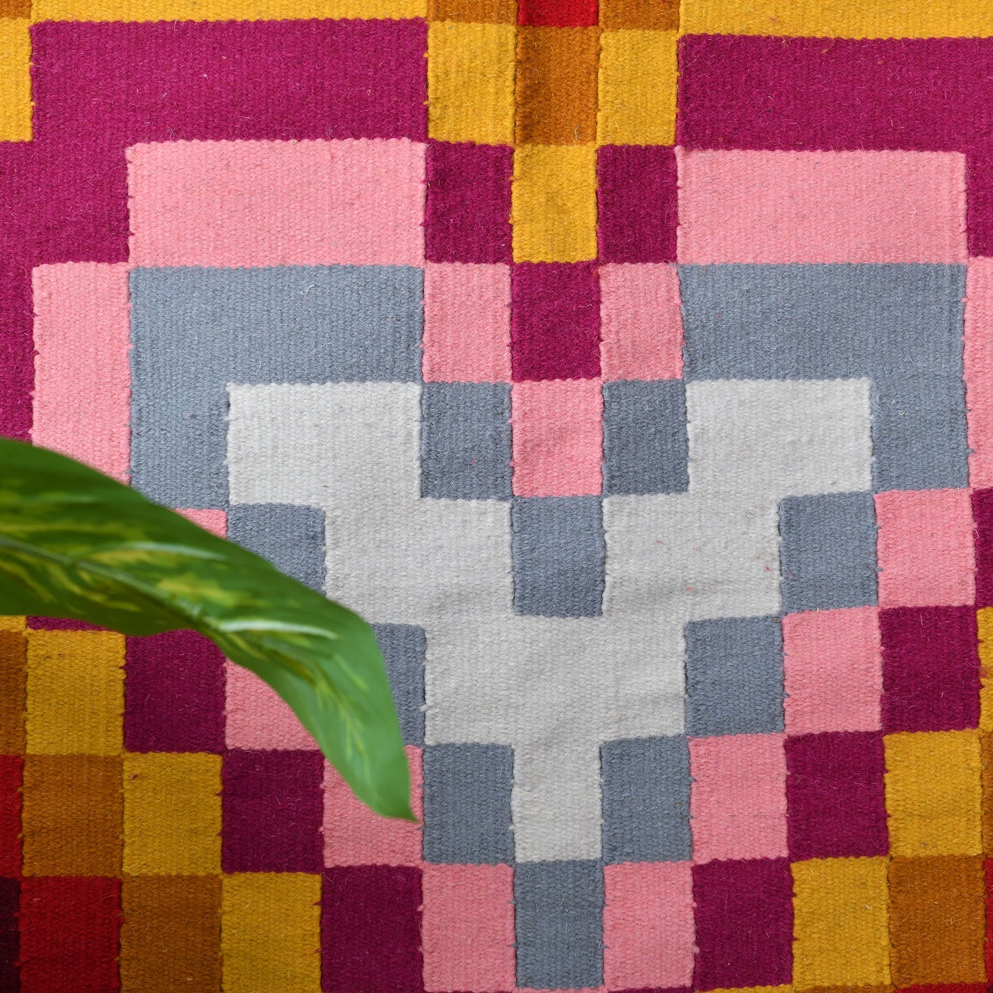 This image shows a close-up of a vibrant geometric heart design woven into a rug. The heart features layers of squares in shades of pink, red, gray, and white. A green plant leaf is partially visible on the left side, adding a natural element to the colorful pattern.