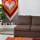 A cozy living room featuring a brown sofa against a white wall. Above the sofa hangs a colorful heart-patterned wall hanging with shades of red, pink, and gray. A vibrant area rug in warm colors lies on the floor, and there is a green plant in the corner.