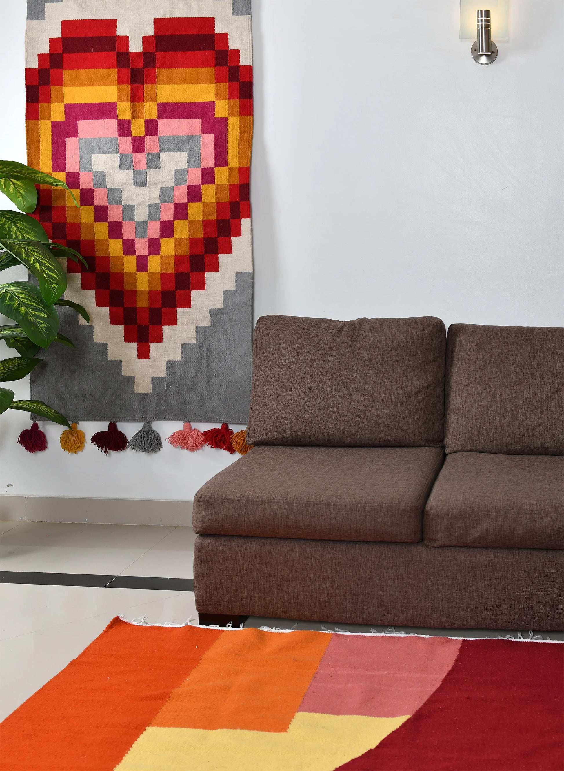 A cozy living room featuring a brown sofa against a white wall. Above the sofa hangs a colorful heart-patterned wall hanging with shades of red, pink, and gray. A vibrant area rug in warm colors lies on the floor, and there is a green plant in the corner.