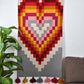 This image shows a colorful woven rug hanging on a wall, featuring a large geometric heart design made up of layered squares in shades of red, pink, orange, and gray. The rug has colorful tassels at the bottom in matching hues. A green plant is positioned on the left, and a brown couch is visible on the right, creating a warm and inviting room setting.