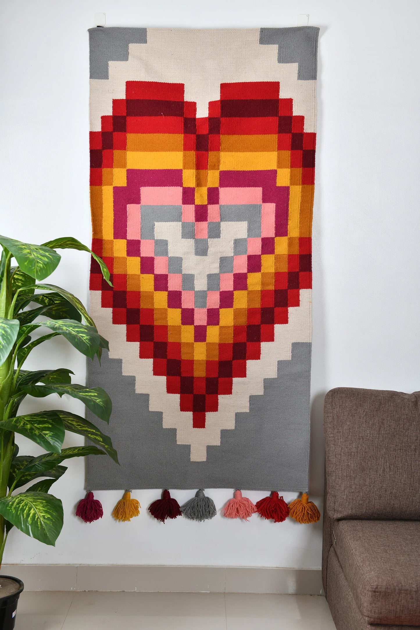 This image shows a colorful woven rug hanging on a wall, featuring a large geometric heart design made up of layered squares in shades of red, pink, orange, and gray. The rug has colorful tassels at the bottom in matching hues. A green plant is positioned on the left, and a brown couch is visible on the right, creating a warm and inviting room setting.