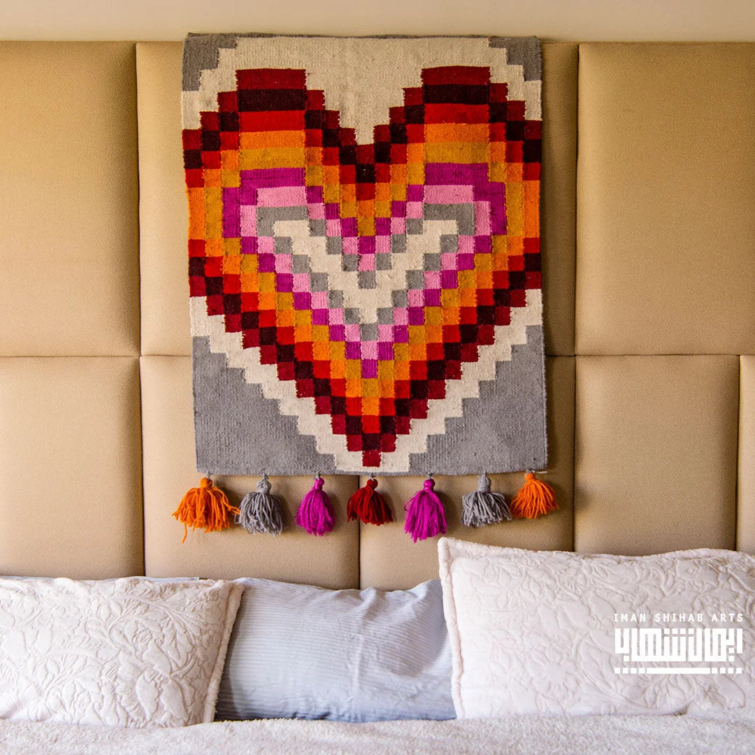 A colorful heart-shaped tapestry in shades of red, pink, and gray, hanging above a light-colored bed with decorative pillows. The tapestry features a geometric design and is adorned with vibrant tassels in purple, orange, and gray.