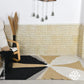 A stylish area rug is displayed on a textured light-colored wall. The rug features a geometric design with a mix of black, beige, white, and grey colors, creating a modern look. Nearby, a wooden chair with a black throw draped over it and decorative elements like dried pampas grass add to the cozy atmosphere.