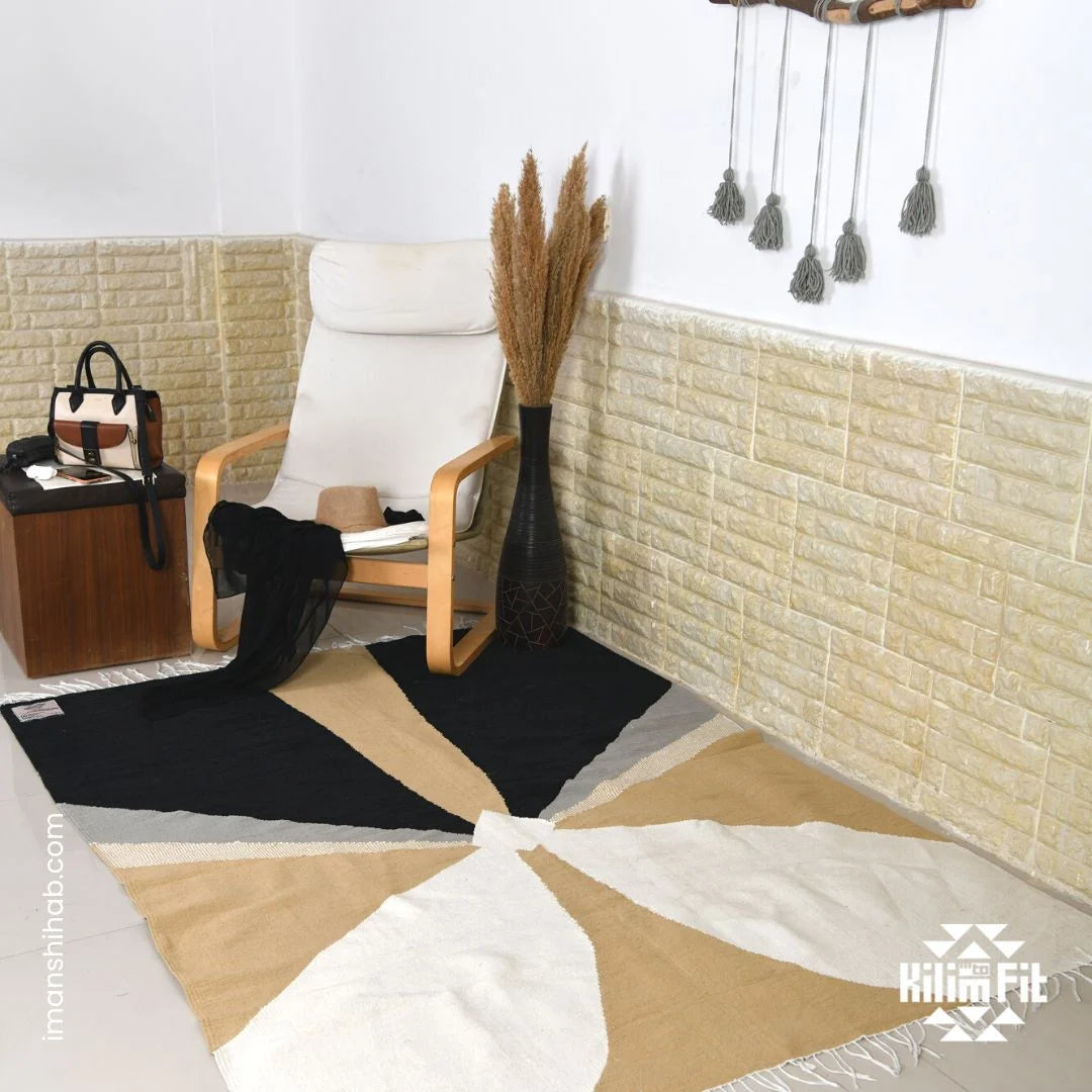 This image shows a cozy corner of a room with a stylish armchair and a geometric rug. The rug features a modern design with sections in black, beige, gray, and white, creating a dynamic pattern. Nearby, a small table holds a handbag and a hat, while decorative dried plants are displayed in the corner, adding warmth to the space.