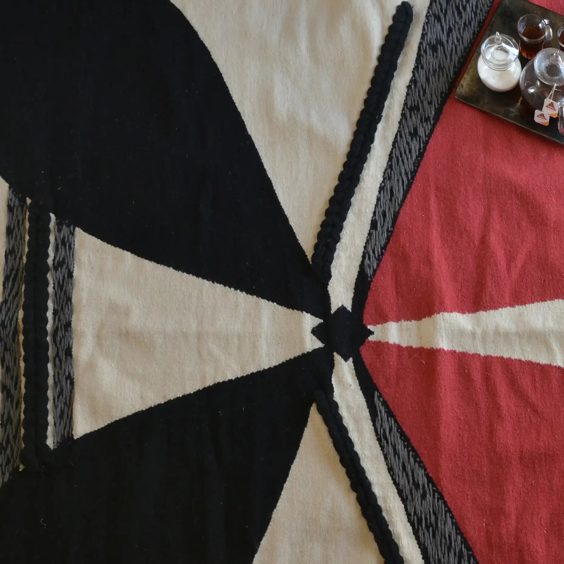 The image features a textile or blanket with a bold geometric design. It includes large sections of black, red, and cream colors, arranged in an abstract pattern with triangular shapes and lines. There are also some small containers or bowls on a tray in the upper right corner, possibly containing spices or other items. The overall composition highlights the contrast between the colors and the intricate design of the fabric.