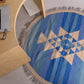 A round area rug with a geometric pattern in various shades of blue and cream, featuring a central star design of triangles. The rug is positioned on a light floor, next to a light wooden desk with a partially visible laptop and a pair of glasses, creating a cozy workspace atmosphere.