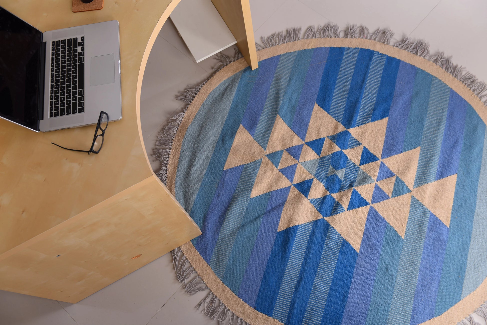 A round area rug with a geometric pattern in various shades of blue and cream, featuring a central star design of triangles. The rug is positioned on a light floor, next to a light wooden desk with a partially visible laptop and a pair of glasses, creating a cozy workspace atmosphere.