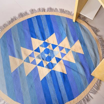 A round area rug featuring a geometric pattern with alternating shades of blue and cream, displaying a central star design made up of triangles. The rug has a fringed edge, and is positioned next to a light-colored wooden table leg, creating a bright and stylish interior setting.