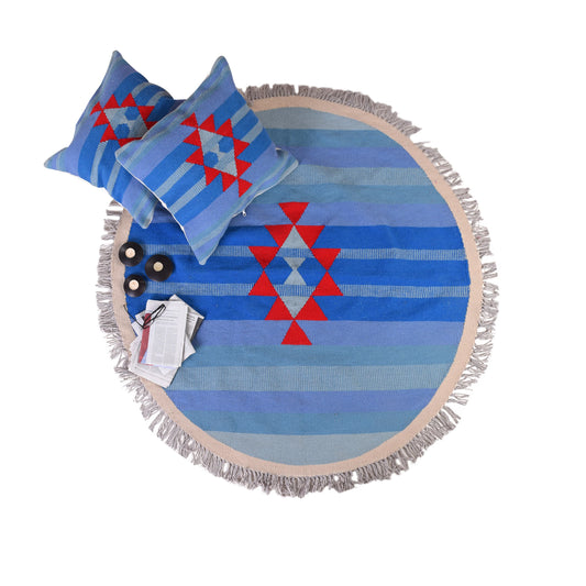 A round, patterned area rug featuring blue and light blue stripes with a central red geometric design. Two matching pillows with a similar pattern are placed on the rug, alongside three black decorative items and a stack of magazines. The rug has a fringed edge, adding texture to the scene.