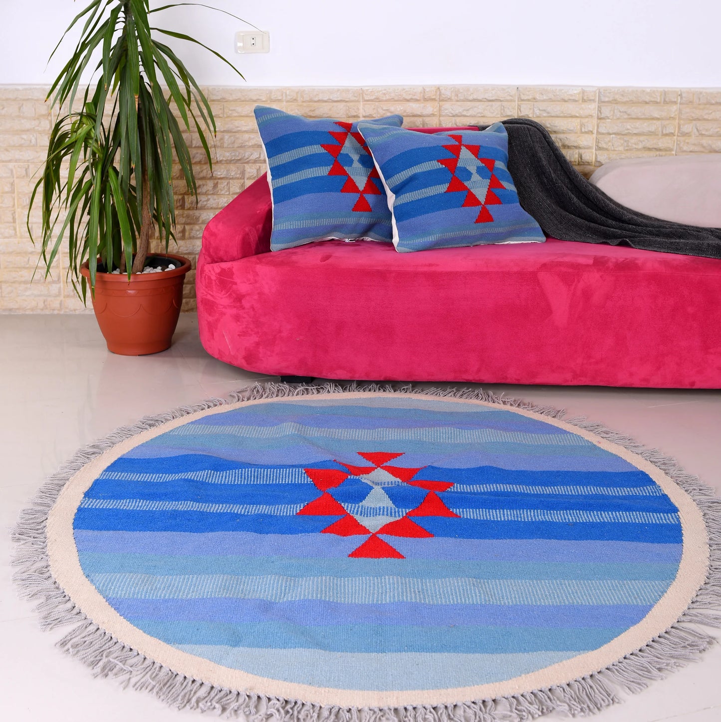 A round area rug featuring a geometric pattern with blue and cream stripes, highlighted by a central red diamond design. The rug is placed on a light floor in front of a pink sofa adorned with two matching decorative pillows. A large potted plant is positioned nearby, enhancing the cozy and vibrant living space.