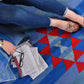  A close-up of a person sitting on a geometric area rug with blue and red patterns. The individual is wearing denim jeans and tan sandals, with one foot resting on the rug. Nearby, a stack of newspapers and a pair of glasses are placed on the rug, creating a relaxed and casual reading environment.