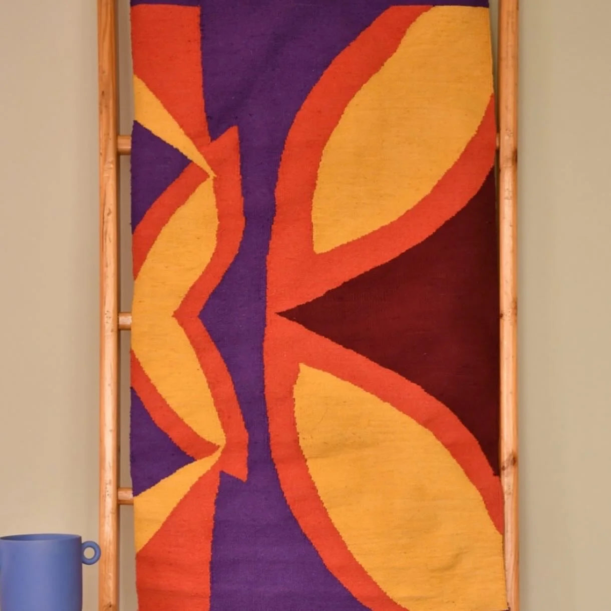 A colorful textile hanging on a wooden ladder, featuring bold geometric patterns in shades of purple, yellow, red, and orange. A blue ceramic jug is positioned at the bottom left of the image.