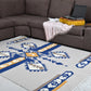 A spacious living area featuring a rectangular area rug with a bold geometric pattern in blue, yellow, and gray. The rug is positioned under a dark brown sectional sofa, where a soft pink throw and a yellow cushion rest. A laptop is placed on the armrest of the sofa, creating a comfortable and functional workspace.