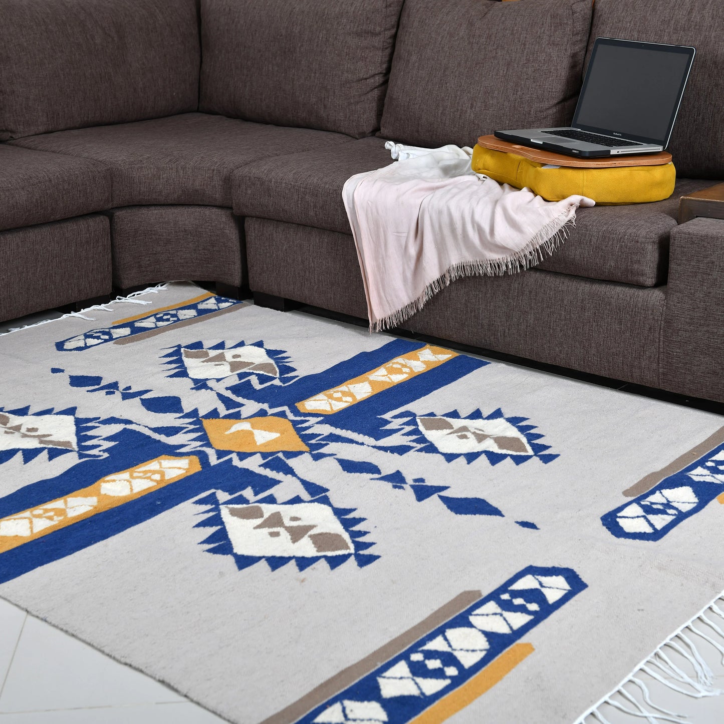 A spacious living area featuring a rectangular area rug with a bold geometric pattern in blue, yellow, and gray. The rug is positioned under a dark brown sectional sofa, where a soft pink throw and a yellow cushion rest. A laptop is placed on the armrest of the sofa, creating a comfortable and functional workspace.