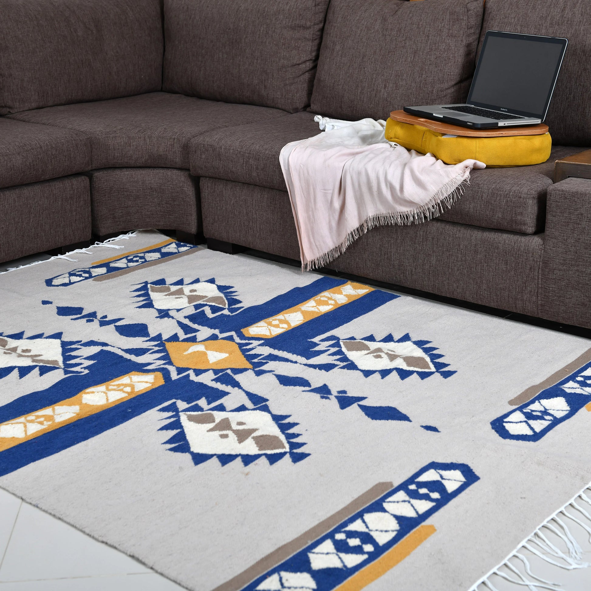A spacious living area featuring a rectangular area rug with a bold geometric pattern in blue, yellow, and gray. The rug is positioned under a dark brown sectional sofa, where a soft pink throw and a yellow cushion rest. A laptop is placed on the armrest of the sofa, creating a comfortable and functional workspace.