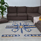 A bright living room featuring a rectangular area rug with a striking geometric design in blue, yellow, and gray. The rug sits beneath a dark brown sectional sofa, complemented by a soft pink throw and a yellow cushion. A lush green plant in a pot adds a touch of greenery to the space, enhancing the inviting atmosphere.
