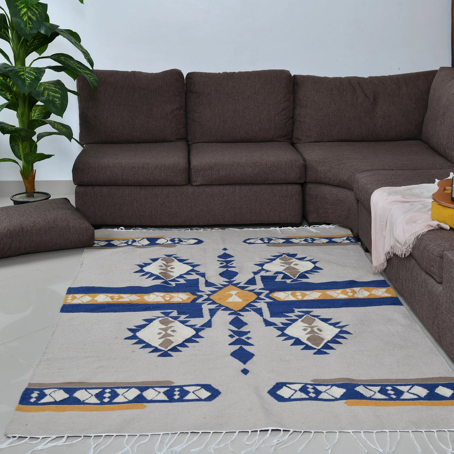 A bright living room featuring a rectangular area rug with a striking geometric design in blue, yellow, and gray. The rug sits beneath a dark brown sectional sofa, complemented by a soft pink throw and a yellow cushion. A lush green plant in a pot adds a touch of greenery to the space, enhancing the inviting atmosphere.