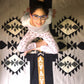 A young girl with glasses sitting cross-legged on a patterned rug, smiling at the camera. She is wearing a light gray long-sleeve shirt and pink pajama pants with a floral design. The rug features a geometric design in black, white, and gold.