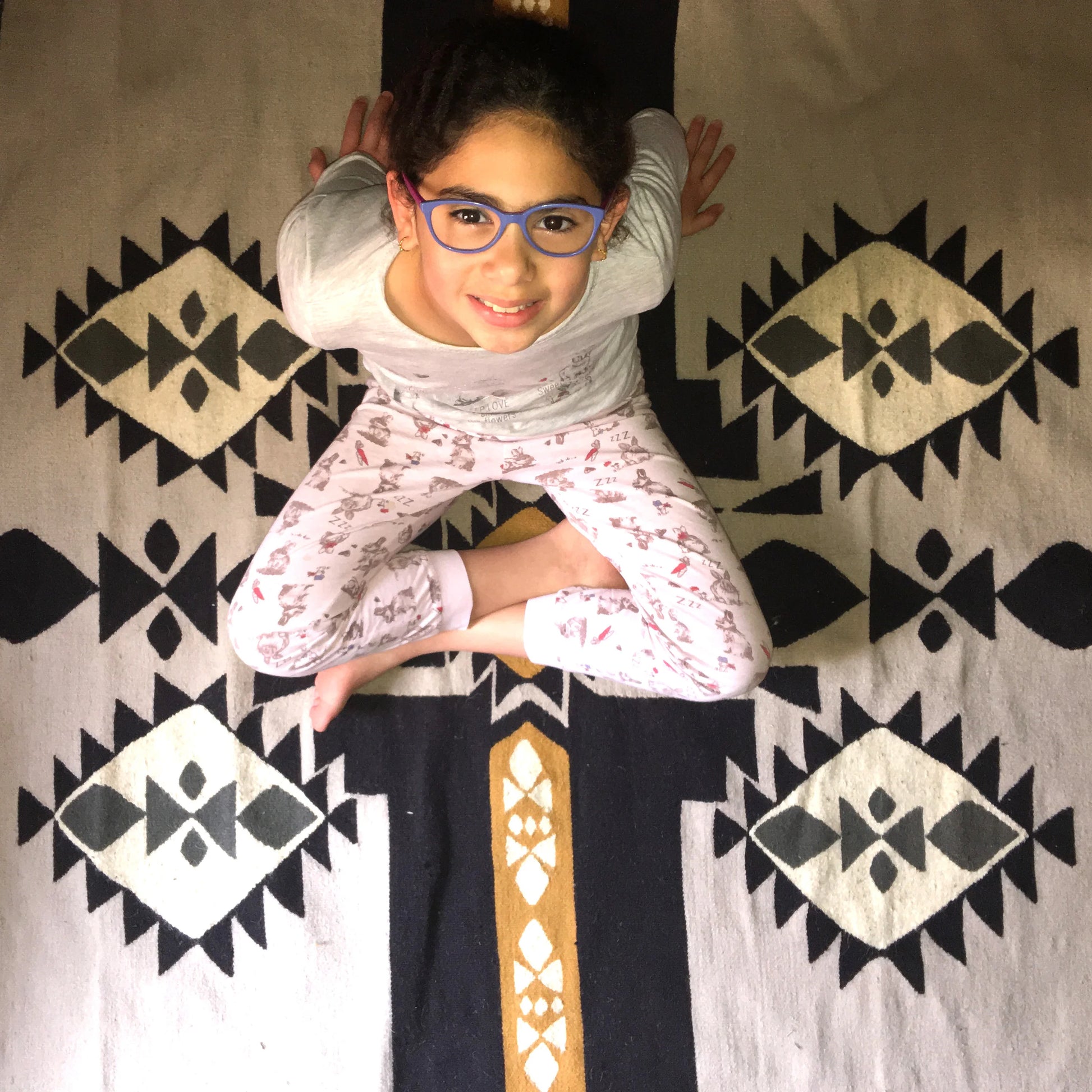 A young girl with glasses sitting cross-legged on a patterned rug, smiling at the camera. She is wearing a light gray long-sleeve shirt and pink pajama pants with a floral design. The rug features a geometric design in black, white, and gold.