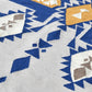 A close-up view of a patterned area rug featuring a geometric design. The rug showcases a mix of blue, yellow, and cream colors, with diamond and triangular shapes interspersed among zigzag patterns. The texture of the fabric is visible, highlighting the intricate details of the design.