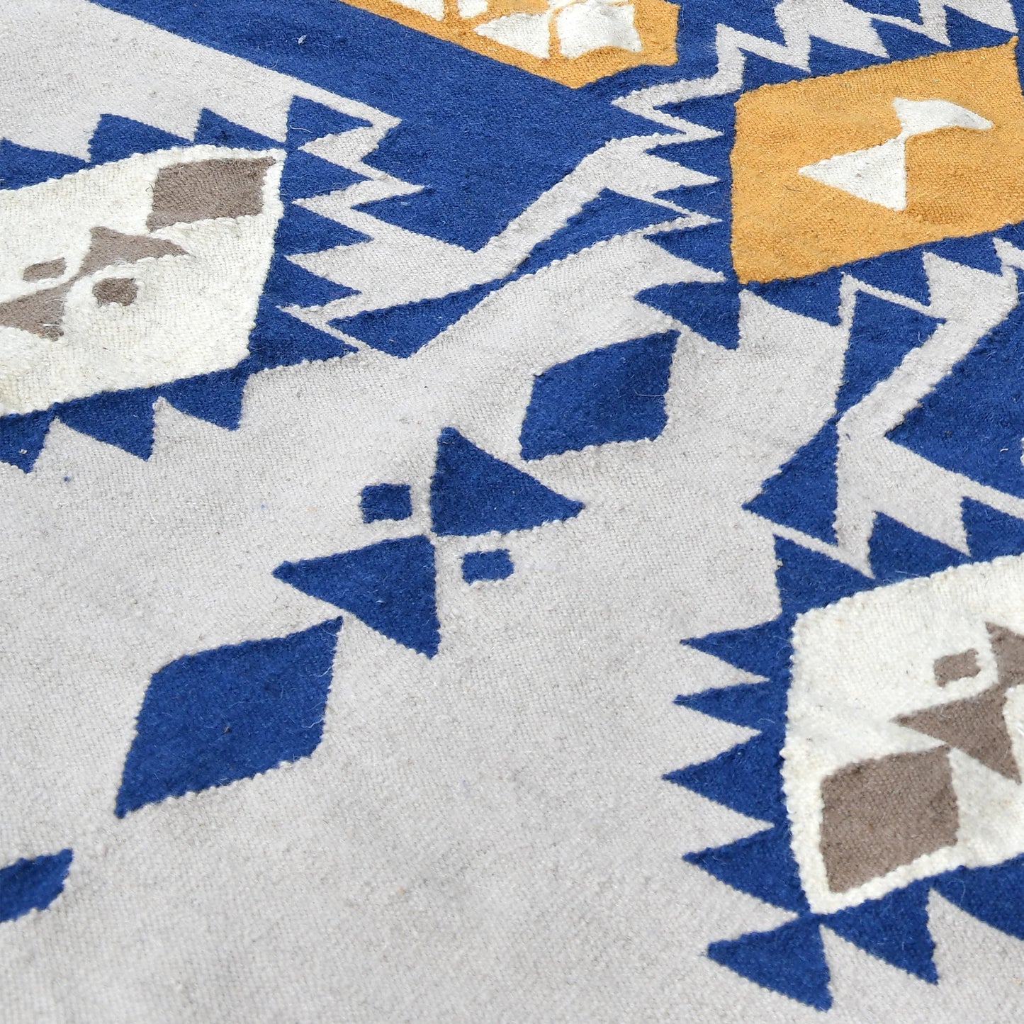 A close-up view of a patterned area rug featuring a geometric design. The rug showcases a mix of blue, yellow, and cream colors, with diamond and triangular shapes interspersed among zigzag patterns. The texture of the fabric is visible, highlighting the intricate details of the design.